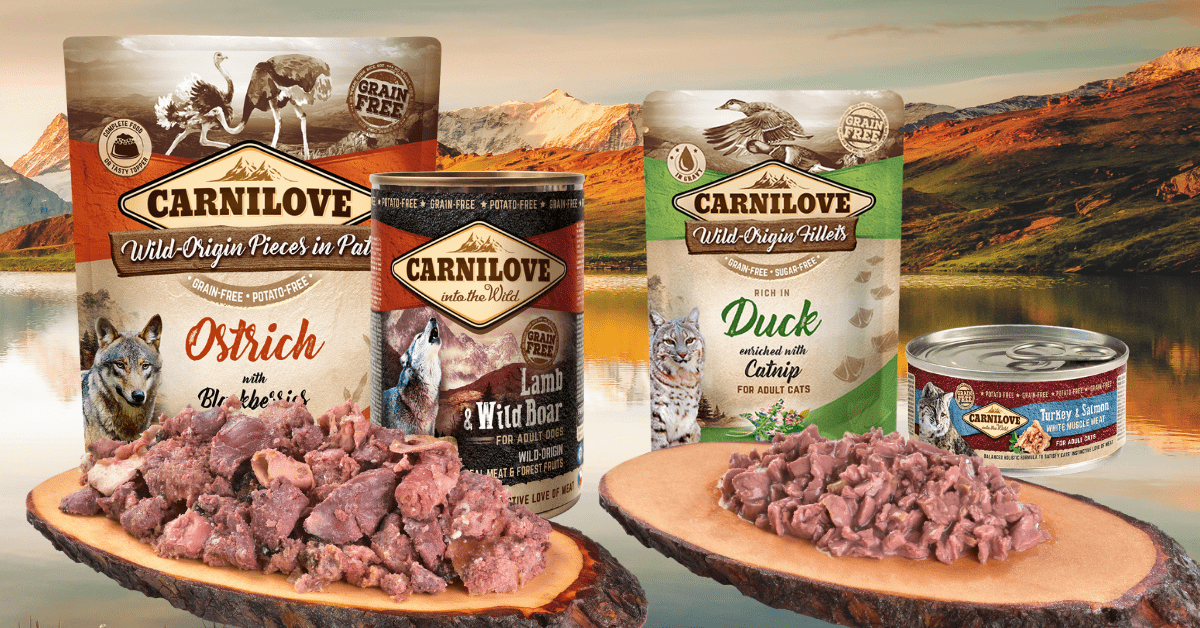 Carnilove food packages on wooden platters, flavours include Ostrich, Lamb, Duck; lake and mountains in background.