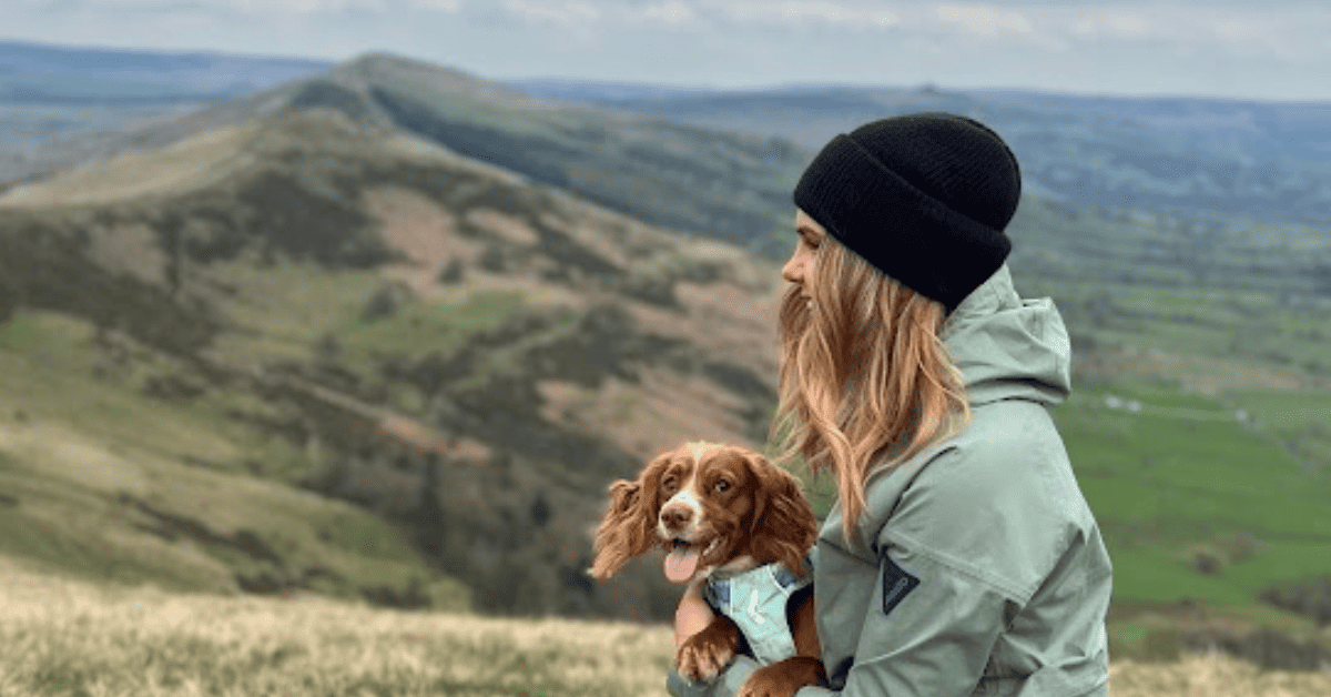 Woman holds small dog, both look at hilly view. She wears beanie and green coat; dog in blue harness. Serene setting.