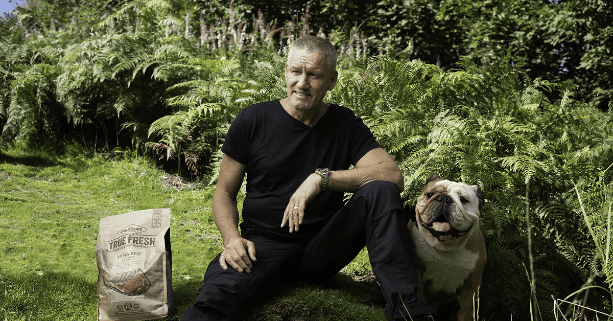 Man in black shirt sits on grass with bulldog. 